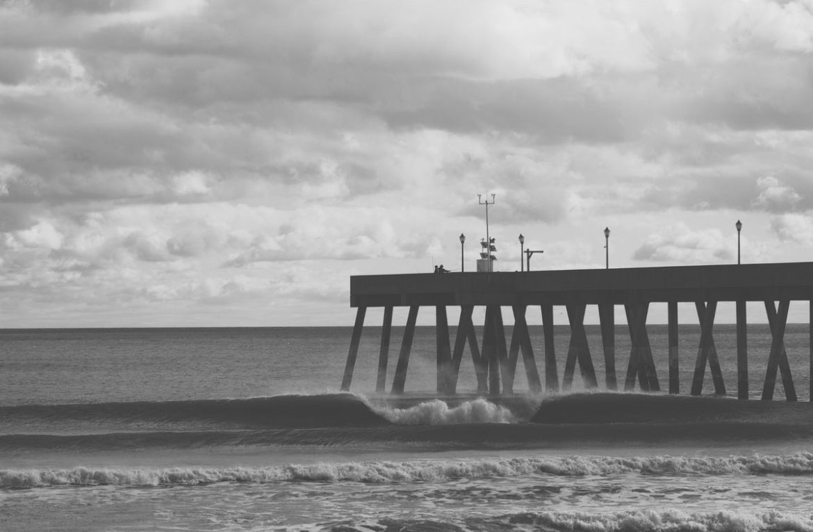 Black and white photograph of a pier extending over ocean waves under a cloudy sky.